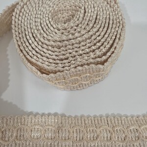 May include: A roll of cream-colored decorative gimp trim featuring a textured, woven surface with a repeating circular pattern along the center. The trim has a scalloped edge and a consistent, uniform width, presented as a coiled spool.