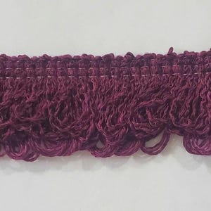 May include: Close-up of a decorative trim in a deep burgundy color. The trim features a woven top edge with a series of looped fringe hanging below. The fringe has a textured, curly appearance, adding a touch of elegance. The trim is against a white background.