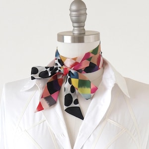 May include: A patterned silk neck scarf in a bow, featuring black polka dots and colourful geometric shapes. The scarf is displayed on a mannequin wearing a white button-down shirt. The scarf's design includes red, pink, yellow, and green squares.