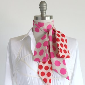 May include: A long, narrow scarf featuring a repeating pattern of pink and red octagonal shapes on a light beige background. The scarf is tied around the neck of a mannequin wearing a crisp white button-down shirt with a structured, crisscross overlay.