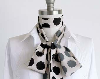 May include: A long, narrow neck scarf featuring a repeating pattern of black and dark grey organic, cloud-like shapes on a light beige background. The scarf is tied in a bow around the neck of a white collared shirt worn by a dress form mannequin.