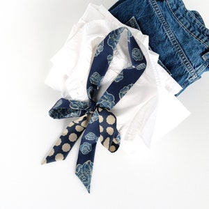 This image showcases a blue rose and dot print skinny scarf lying on a casually stacked white shirt and pair of blue jeans. The blue shades of the scarf tie the elements of the image together harmoniously.