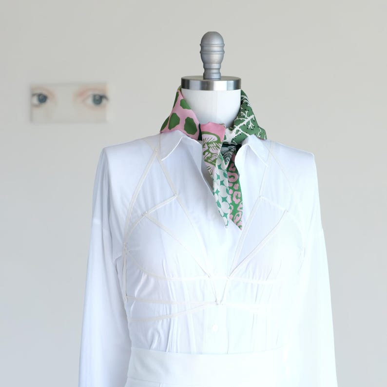 May include: A white long-sleeve shirt with a colorful scarf. The scarf features pink and green patterns, including polka dots and floral designs. The shirt is displayed on a mannequin.