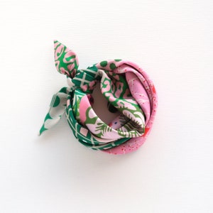 May include: A rolled silk scarf with a vibrant floral pattern in shades of pink, green, and white. The scarf is knotted at one end, showcasing the colorful design. The scarf is on a white background.