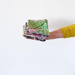 May include: A stack of colorful fabric squares held in a person's hand. The fabrics feature various patterns, including floral, geometric, and abstract designs. The color palette includes pink, green, blue, and yellow. The fabrics appear to be folded and ready for use in a sewing or craft project.
