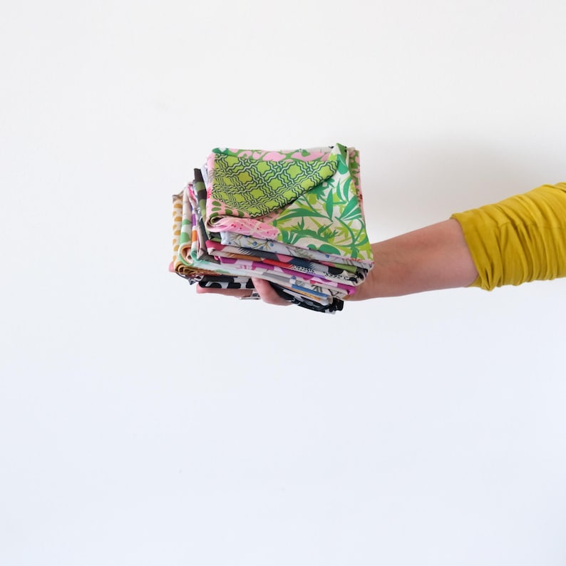 May include: A stack of colorful fabric squares held in a person's hand. The fabrics feature various patterns, including floral, geometric, and abstract designs. The color palette includes pink, green, blue, and yellow. The fabrics appear to be folded and ready for use in a sewing or craft project.