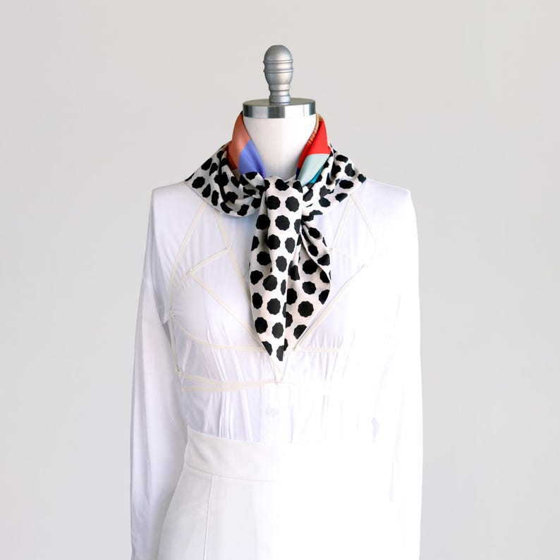 Puede incluir: Una camisa blanca con una bufanda de lunares blancos y negros atada alrededor del cuello. La bufanda tiene un patr&oacute;n geom&eacute;trico colorido en la parte posterior.