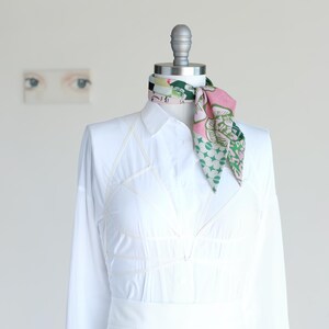 A pretty pink and green patchwork print small triangle scarf is rolled and tied as a neckerchief on a mannequin in a white shirt dress.