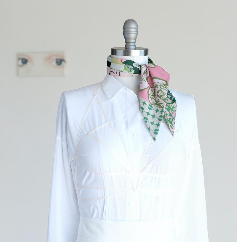 A pretty pink and green patchwork print small triangle scarf is rolled and tied as a neckerchief on a mannequin in a white shirt dress.