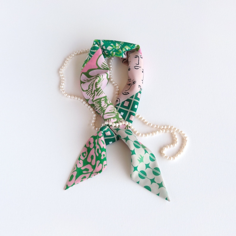 A pretty pink and green patchwork print small triangle scarf styled with a strand of pearls in a flat-lay photo.