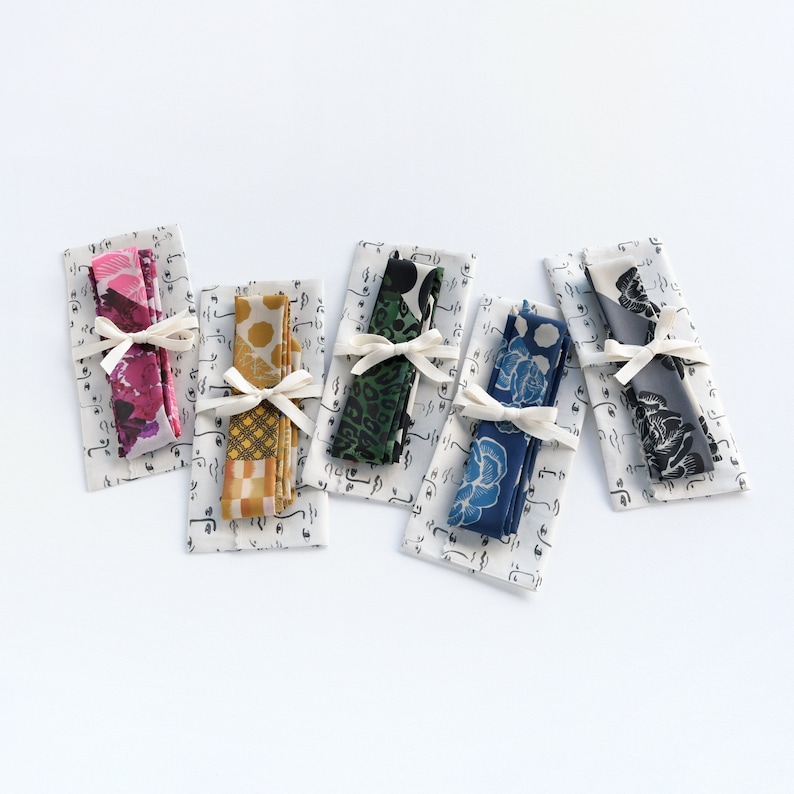 The image portrays a collection of five different colored skinny scarves folded and sitting on protective pouches and tied with a ribbon. Colors from left to right; burgundy/red, yellow/gold, emerald green/black, blue/off-white, and gray/black.