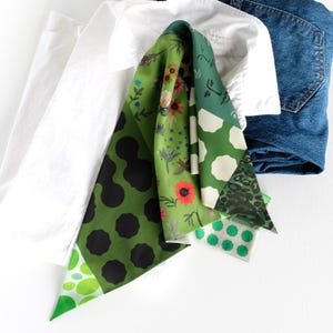 K&ouml;nnte beinhalten: Ein gemusterter Schal in Gr&uuml;n- und Wei&szlig;t&ouml;nen mit schwarzen Punkten, floralen Motiven und Tupfen. Der Schal liegt &uuml;ber einem wei&szlig;en Hemd und einer blauen Jeans.