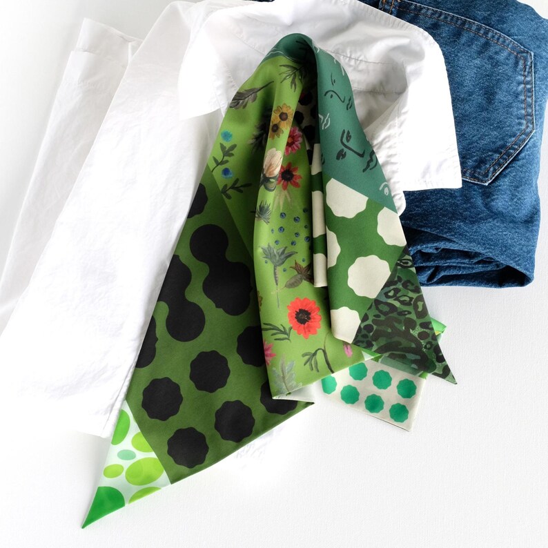 K&ouml;nnte beinhalten: Ein gemusterter Schal in Gr&uuml;n- und Wei&szlig;t&ouml;nen mit schwarzen Punkten, floralen Motiven und Tupfen. Der Schal liegt &uuml;ber einem wei&szlig;en Hemd und einer blauen Jeans.