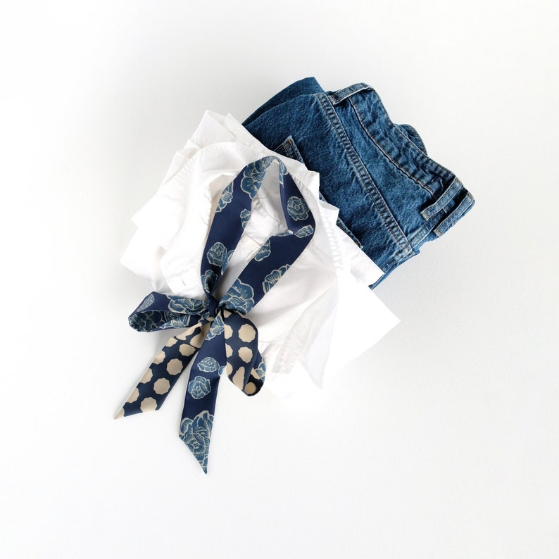 This image showcases a blue rose and dot print skinny scarf lying on a casually stacked white shirt and pair of blue jeans. The blue shades of the scarf tie the elements of the image together harmoniously.