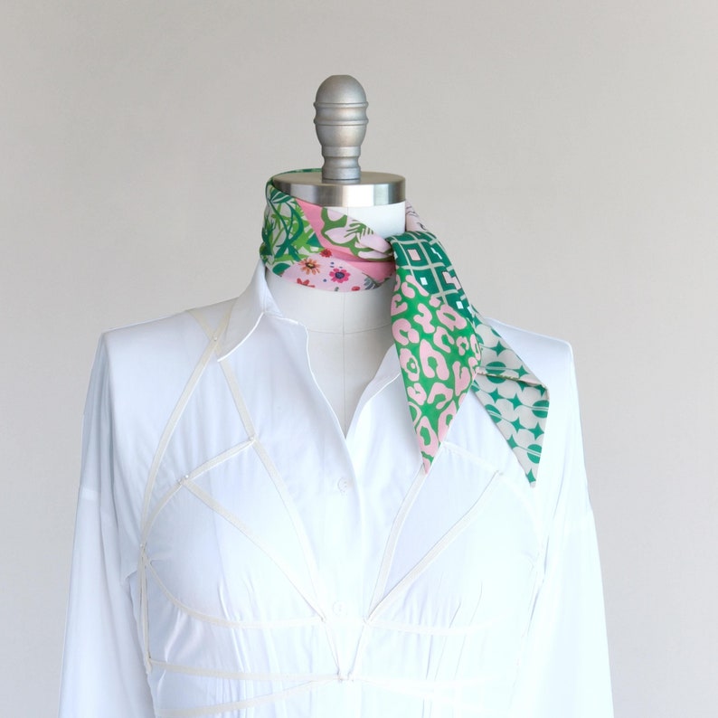 A pretty pink and green patchwork print small triangle scarf is rolled and tied as a neckerchief on a mannequin in a white shirt dress.