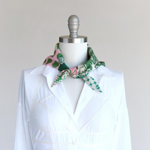 A pretty pink and green patchwork print small triangle scarf tied in a sailor style with a knot at the neck front on a mannequin in a white shirt dress.