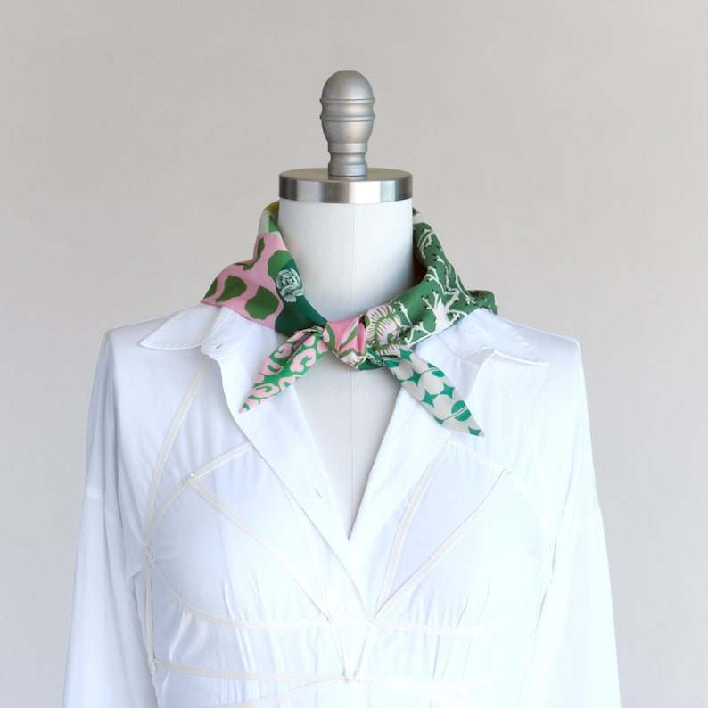 A pretty pink and green patchwork print small triangle scarf tied in a sailor style with a knot at the neck front on a mannequin in a white shirt dress.