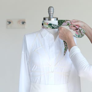 May include: A white long-sleeved shirt on a mannequin with a colorful floral scarf being tied around the neck. The shirt has a unique design with visible seams. The background is a plain white wall with a small painting of eyes.