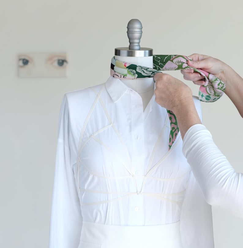 May include: A white long-sleeved shirt on a mannequin with a colorful floral scarf being tied around the neck. The shirt has a unique design with visible seams. The background is a plain white wall with a small painting of eyes.