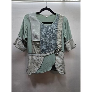 May include: A sage green short-sleeved top featuring a patchwork design with various floral patterns, textured fabric panels, and a small fabric rose accent at the waist. The garment has a round neckline and raw-edge seams throughout the construction.