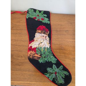 May include: A black needlepoint Christmas stocking featuring a central image of Santa Claus with a white beard and red hat. The stocking is decorated with green holly leaves and red berries, finished with a red fabric trim and a red hanging loop.
