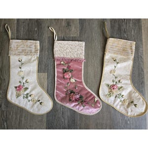 Puede incluir: Tres calcetines decorativos con lazos para colgar. Dos calcetines son de color crema con un sutil patrón de damasco, puños plisados y enredaderas florales bordadas. El calcetín central es de terciopelo malva con un puño estampado y rosas.