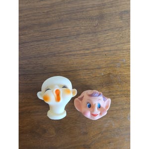 May include: Two small molded plastic doll heads. One head is pale white with closed eyes, long black eyelashes, orange cheeks, and an open orange mouth. The second head is peach-colored with pointed ears, blue eyes, brown hair, and a red mouth.