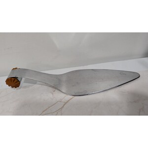 May include: A silver-toned metal cake server with a pointed, flat blade and a curved handle. The end of the handle features a small, round, brown plastic accent with a ridged, star-like pattern on its surface, providing a decorative detail.