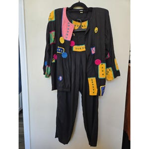 May include: A black two-piece outfit consisting of a long-sleeved jacket and matching trousers. The jacket features a pink lapel and various colorful patches in yellow, green, blue, and magenta, accented with small circular studs throughout the garment.