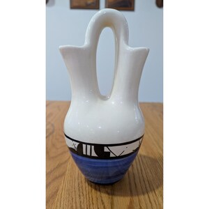 May include: A cream-colored ceramic wedding vase featuring a double-spout design with a central arched handle. The lower portion of the vessel is decorated with a horizontal band of black geometric patterns above a solid blue base section.