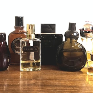 May include: A collection of eight glass perfume bottles of varying shapes and sizes, including a rectangular Aramis bottle, a dark brown round bottle, a tall amber bottle labeled Soul, a black square bottle, and a small clear glass miniature bottle.