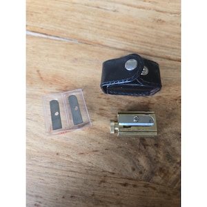 Pode incluir: Um apontador de latão com botão de ajuste serrilhado, um pequeno estojo de couro preto com fecho de botão de pressão prateado e um recipiente de plástico transparente contendo duas lâminas de reposição de metal.