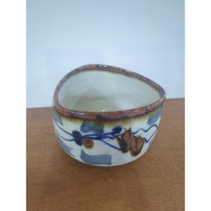 May include: A ceramic bowl with an irregular, rounded triangular rim. The exterior features a cream-coloured base with hand-painted blue swirling lines, brown leaf-like shapes, and grey patches. The rim is accented with a dark brown glaze.