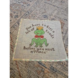 May include: A square needlepoint canvas featuring a green frog wearing a yellow crown and a red bow tie. The text reads: You have to kiss a lot of toads before you meet a Prince. The canvas has a grid pattern and a white border with green text.