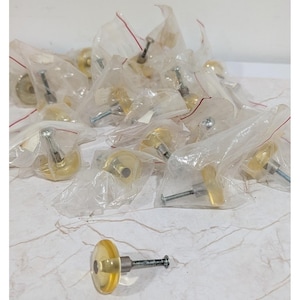 Puede incluir: Una colección de pomos para armarios redondos y translúcidos de color amarillo, cada uno con una base de metal plateado y un tornillo roscado. Varios de estos pomos están empaquetados individualmente en bolsas de plástico transparente.
