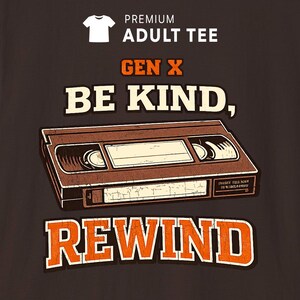 May include: A dark brown t-shirt featuring a graphic of a vintage VHS cassette tape. The text reads "PREMIUM ADULT TEE", "GEN X", "BE KIND,", and "REWIND" in a distressed, retro-style font with orange and cream colours against the dark background.