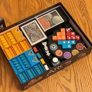May include: A board game box with various components, including cards, dice, and tokens. The box is divided into compartments with yellow, blue, and orange pieces. The game's title, "ARCS", is visible on the box.