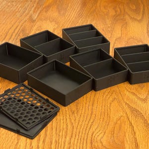 May include: A collection of black plastic storage containers, some with dividers, and a separate black lid with a honeycomb pattern. The containers are arranged on a wooden surface, suggesting an organizational theme.