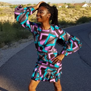 May include: A woman wearing a black, purple, and turquoise dress with a geometric pattern. The dress has long sleeves and a ruffled hem.