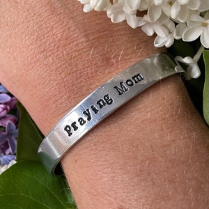 May include: A silver-colored metal cuff bracelet with the stamped text "Praying Mom" is displayed. The bracelet is worn on a wrist, surrounded by white and purple flowers.