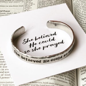 May include: A silver-toned cuff bracelet with the stamped phrase "She believed He could so she prayed." The bracelet is set on a white surface with text from a book in the background. The bracelet has a hammered texture.