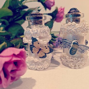 Könnte beinhalten: Zwei dekorative Glasflaschen mit Korken und Kristallaufsätzen. Eine Flasche hat einen Schmetterling, die andere eine Biene. Beide sind mit weißer Spitze, Perlen und Glitzer verziert. Rosa und lila Blumen sind im Hintergrund.