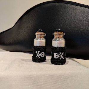 May include: Two small glass bottles filled with glitter, each adorned with a black skull and crossbones design. The bottles have cork stoppers and sit in front of a black pirate hat.