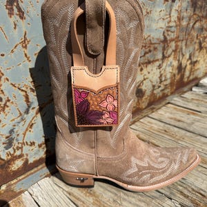 Könnte beinhalten: Ein brauner Wildleder-Cowboystiefel mit einer floralen Ledertasche. Die Tasche ist mit rosa und lila Blumen und einem Wabenmuster verziert. Der Stiefel hat dekorative Nähte und eine spitze Zehenpartie.