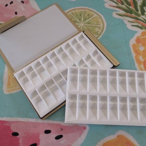 Puede incluir: Una paleta de plástico blanca con 24 compartimentos, ideal para guardar y organizar objetos pequeños. La paleta está en una caja de metal dorado con una tapa de espejo.