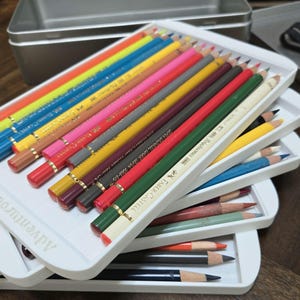 Puede incluir: Un juego de lápices de colores Faber-Castell Polychromos dispuestos en bandejas de plástico blanco. Los lápices presentan una variedad de tonos vibrantes que incluyen rojo, amarillo, azul, verde y marrón. La bandeja blanca tiene el texto Adventurous Art.