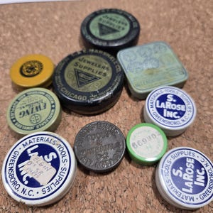 Puede incluir: Una colección de latas antiguas de suministros para relojes y joyas en varias formas y colores. Las latas presentan textos como "Jewelers Supplies" y "Materials Tools Supplies". Algunas latas tienen etiquetas escritas a mano.