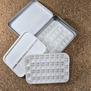 May include: A white rectangular paint palette with a lid and a separate tray, alongside a metal tin. The palette has multiple small square compartments. The lid and tray are white, while the tin has a silver rim.