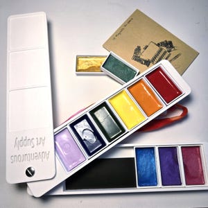 Pode incluir: Um conjunto de tintas de aquarela em uma caixa de plástico branca. A caixa tem um rótulo que diz "Adventurous Art Supply". As tintas são organizadas em filas de quatro, com cada fila contendo uma cor diferente. As cores são roxo, azul, verde, amarelo, laranja, vermelho, azul, roxo e rosa.