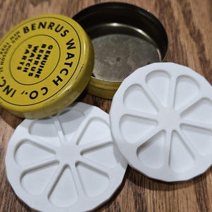 May include: A yellow metal tin with the text "BENRUS WATCH CO." and "GENUINE BENRUS WATCH PARTS" in black. Two white, circular watch parts are next to the tin. The tin is open, revealing a silver interior.